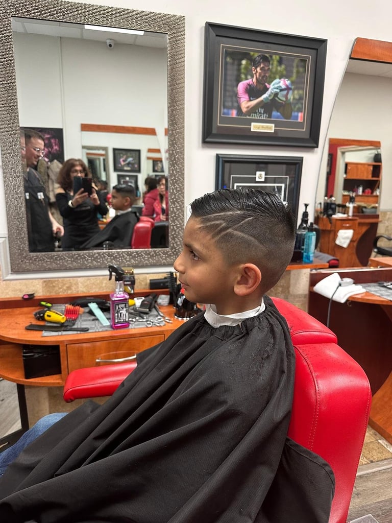 Young boy getting a haircut in a barber shop chair, wearing a black cape, with mirror and framed photos visible