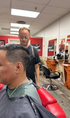 Barber smiling at camera while cutting customers hair in a salon chair, with red and wood interior visible