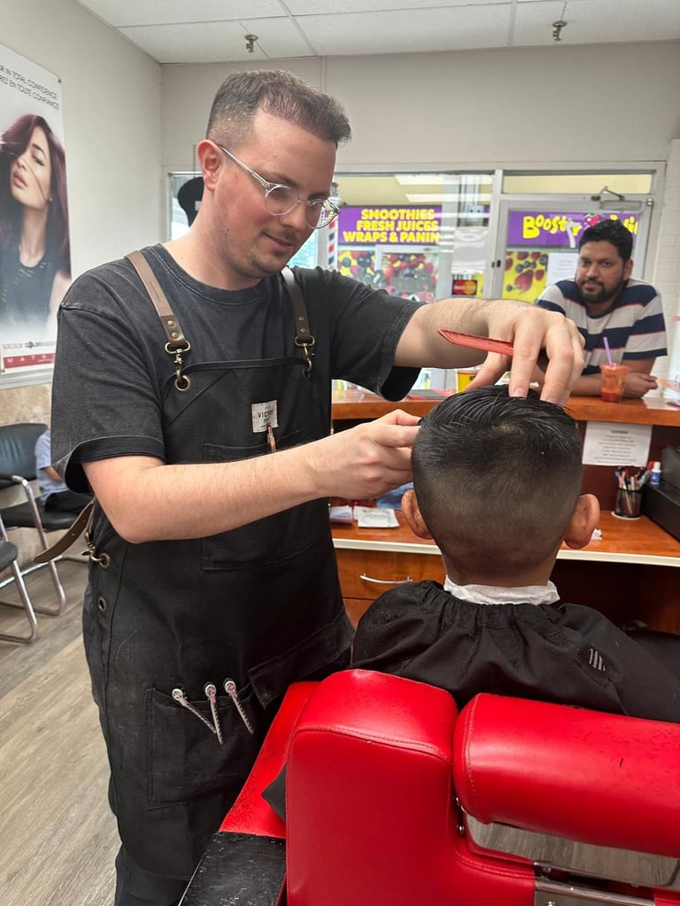 Barber in black apron giving a haircut to a young customer in a salon chair with colorful posters in background