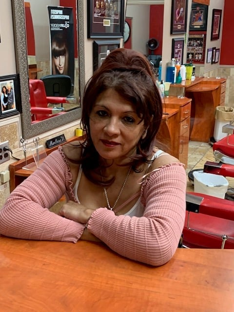 Woman with brown hair wearing a pink top poses at a salon counter with mirrors and styling products in the background