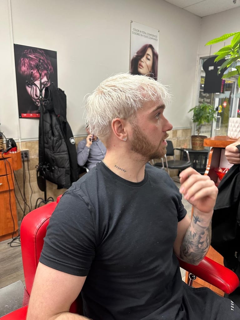 Man with short white hair and tattoo on arm sits in salon chair facing left, with posters and plants visible in background
